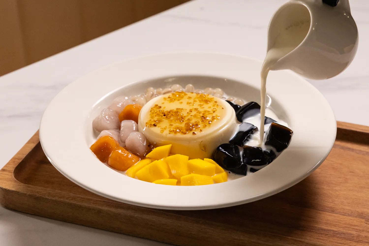 A white bowl of Taiwanese-style dessert featuring mango chunks, taro balls, black grass jelly, and a central custard pudding topped with golden syrup, as milk is being poured over it.